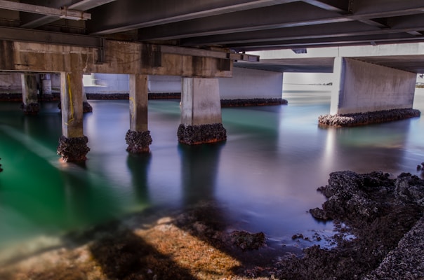 A view under a bridge with concrete pillars extending into a body of water. The water appears calm and reflects a gradient of green and blue hues. Barnacles and seaweed are attached to the lower parts of the pillars and the rocky shoreline.