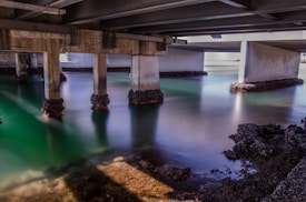 A view under a bridge with concrete pillars extending into a body of water. The water appears calm and reflects a gradient of green and blue hues. Barnacles and seaweed are attached to the lower parts of the pillars and the rocky shoreline.
