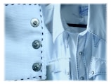 A close-up of a white cowboy-style shirt with contrasting black stitching. The fabric has a textured, ribbed appearance. The shirt features metal snap buttons, with two visible on a button placket and several more on the pockets and collar. A clothing label is seen on the inside of the shirt's collar.