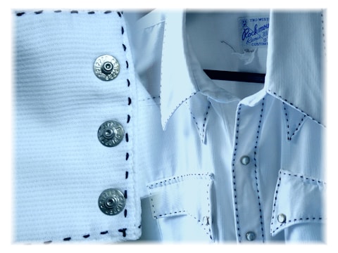 A close-up of a white cowboy-style shirt with contrasting black stitching. The fabric has a textured, ribbed appearance. The shirt features metal snap buttons, with two visible on a button placket and several more on the pockets and collar. A clothing label is seen on the inside of the shirt's collar.