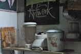 A kitchen shelf holds a few appliances and utensils. There is a blender with its jar placed next to a traditional kettle labeled 'HOT.' A glass beverage pitcher is present, alongside a bamboo drying rack for utensils. Above, a chalkboard with artistic lettering reads 'Artwork.' The setting has an eclectic and vintage aesthetic, partly due to the kitchen’s aged decor and utensils.