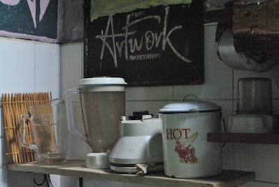A kitchen shelf holds a few appliances and utensils. There is a blender with its jar placed next to a traditional kettle labeled 'HOT.' A glass beverage pitcher is present, alongside a bamboo drying rack for utensils. Above, a chalkboard with artistic lettering reads 'Artwork.' The setting has an eclectic and vintage aesthetic, partly due to the kitchen’s aged decor and utensils.