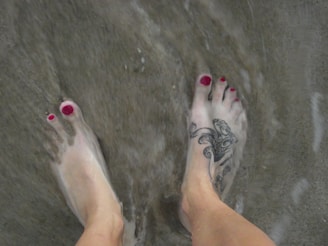 Two feet with red-painted toenails are partly submerged in shallow water, with sand visible underneath. One foot has a tattoo featuring a decorative design.