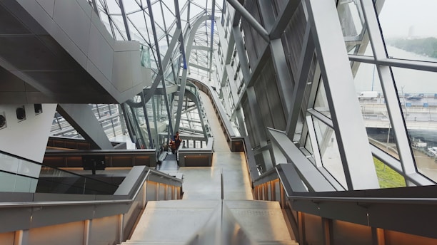 A modern architectural interior featuring large metal beams, expansive glass windows, and an angled ceiling. The space has multiple levels connected by staircases, escalators, and walkways. The area appears spacious and open, with minimalistic design elements.