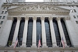 The image features the facade of a grand building with Corinthian columns and a pediment adorned with sculptures. The building is identified as the New York Stock Exchange, with its name prominently displayed. American flags are visible in front of the structure, adding to its formal and historic appearance.