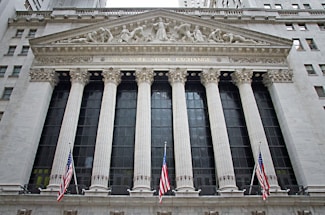 The image features the facade of a grand building with Corinthian columns and a pediment adorned with sculptures. The building is identified as the New York Stock Exchange, with its name prominently displayed. American flags are visible in front of the structure, adding to its formal and historic appearance.