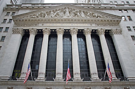 The image features the facade of a grand building with Corinthian columns and a pediment adorned with sculptures. The building is identified as the New York Stock Exchange, with its name prominently displayed. American flags are visible in front of the structure, adding to its formal and historic appearance.