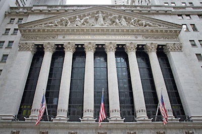 The image features the facade of a grand building with Corinthian columns and a pediment adorned with sculptures. The building is identified as the New York Stock Exchange, with its name prominently displayed. American flags are visible in front of the structure, adding to its formal and historic appearance.