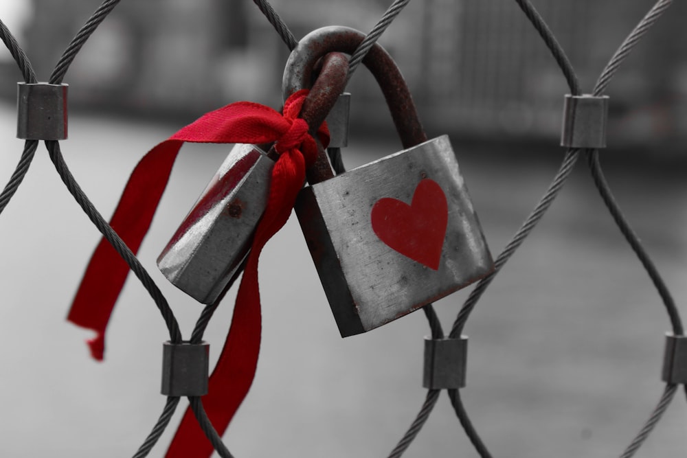 Padlock on a wire fence has red heart painted on it and ribbon attached