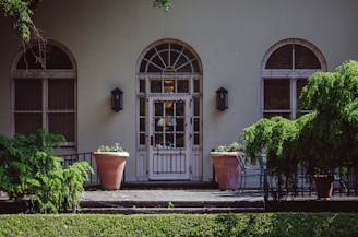 A building facade featuring three large arched windows with decorative metal grilles on either side of a central door. The entrance is flanked by two large terracotta planters with green plants. There are two wall lanterns on either side of the door, and lush green bushes partially obscure the view.