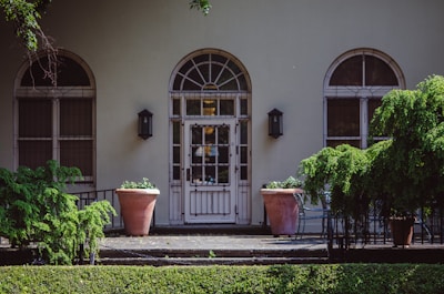 A building facade featuring three large arched windows with decorative metal grilles on either side of a central door. The entrance is flanked by two large terracotta planters with green plants. There are two wall lanterns on either side of the door, and lush green bushes partially obscure the view.