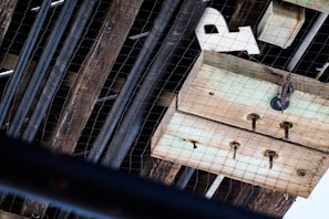A wooden structure is visible with several old, rusted nails and metal components attached to it. There are thick beams running parallel, with a wire mesh covering part of the scene. Some objects, possibly wooden panels or boxes, are partially obstructed by the netting. The angle suggests an overhead view, possibly taken from below.