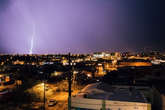 A nighttime urban scene with a prominent bolt of lightning illuminating the sky. Buildings and streets are visible below, with city lights scattered across the area. The sky is a mixture of dark and bright hues due to the lightning.
