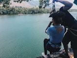 Two individuals preparing for a zip line adventure over a river, surrounded by dense vegetation and mountains in the background. The person in the foreground is wearing a harness and appears ready to launch off the edge while being assisted by another person.