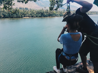 Two individuals preparing for a zip line adventure over a river, surrounded by dense vegetation and mountains in the background. The person in the foreground is wearing a harness and appears ready to launch off the edge while being assisted by another person.