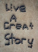 Letters formed with small, round stones on a sandy-colored surface spell out the phrase 'Live A Great Story'.