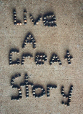 Letters formed with small, round stones on a sandy-colored surface spell out the phrase 'Live A Great Story'.