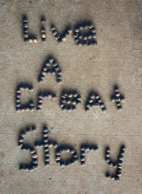 Letters formed with small, round stones on a sandy-colored surface spell out the phrase 'Live A Great Story'.