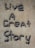 Letters formed with small, round stones on a sandy-colored surface spell out the phrase 'Live A Great Story'.