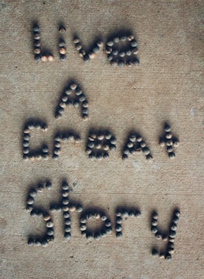 Letters formed with small, round stones on a sandy-colored surface spell out the phrase 'Live A Great Story'.