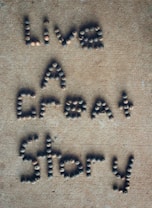 Letters formed with small, round stones on a sandy-colored surface spell out the phrase 'Live A Great Story'.