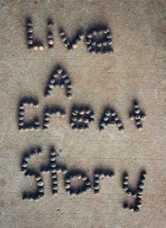 Letters formed with small, round stones on a sandy-colored surface spell out the phrase 'Live A Great Story'.