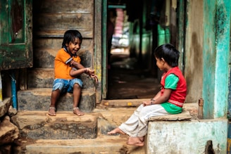 Two young children are sitting on a set of worn stone steps, facing each other. The child on the left is wearing an orange shirt and denim shorts, while the child on the right is dressed in a red vest and green shirt with light-colored pants. Both appear to be engaged in a playful conversation or game. The surroundings are rustic, with the stone steps leading into a dimly lit interior space.