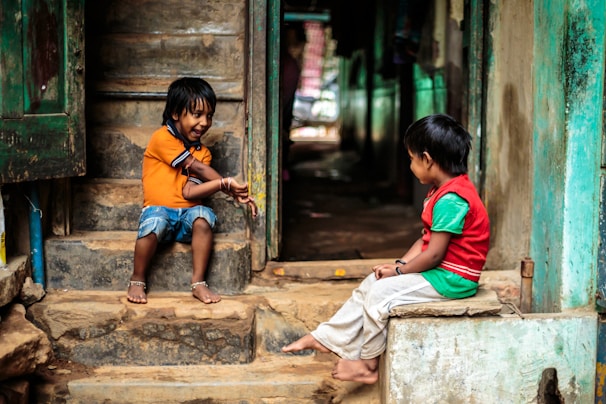 Two young children are sitting on a set of worn stone steps, facing each other. The child on the left is wearing an orange shirt and denim shorts, while the child on the right is dressed in a red vest and green shirt with light-colored pants. Both appear to be engaged in a playful conversation or game. The surroundings are rustic, with the stone steps leading into a dimly lit interior space.