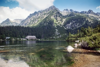A picturesque mountain landscape features a large alpine lake surrounded by dense evergreen forests. In the background, rugged mountains rise sharply with rocky peaks visible against a blue sky dotted with fluffy clouds. A lodge-like building with a steep roof is nestled among the trees near the water's edge, suggesting a tranquil retreat. The clear water of the lake reflects the surrounding scenery, adding to the serene atmosphere.