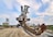 A massive industrial excavator with a large bucket-wheel is situated in a desert-like landscape. The machine features a series of gears and metallic components, appearing heavy and robust. Its structure is extensive, with conveyors and other mining equipment visible. In the background, green trees line the horizon under a partly cloudy sky.