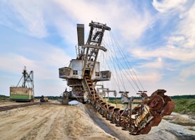 A massive industrial excavator with a large bucket-wheel is situated in a desert-like landscape. The machine features a series of gears and metallic components, appearing heavy and robust. Its structure is extensive, with conveyors and other mining equipment visible. In the background, green trees line the horizon under a partly cloudy sky.