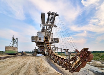 A massive industrial excavator with a large bucket-wheel is situated in a desert-like landscape. The machine features a series of gears and metallic components, appearing heavy and robust. Its structure is extensive, with conveyors and other mining equipment visible. In the background, green trees line the horizon under a partly cloudy sky.