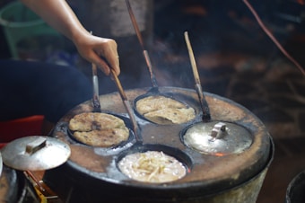 A hand is using a tool to flip flatbreads on a round, multi-sectioned stove. The stove has circular moulds where flatbreads are cooking, and steam rises from the surface. The setting appears to be an outdoor or street food scene.