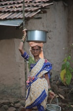 An elderly woman is dressed in traditional colorful attire and is carrying a metal pot on her head. She holds a bucket in her left hand. She stands in front of an old, rustic building with tiled roofing. The background includes simple, natural surroundings.