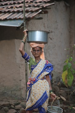 An elderly woman is dressed in traditional colorful attire and is carrying a metal pot on her head. She holds a bucket in her left hand. She stands in front of an old, rustic building with tiled roofing. The background includes simple, natural surroundings.