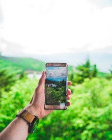 A hand is holding a smartphone with a scenic landscape displayed on its screen. The background consists of lush green trees and a bright sky. The smartphone's screen shows a similar landscape, creating a blend between the physical and digital worlds.