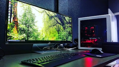 A sleek computer setup featuring a large monitor displaying a forest scene with vibrant greenery. Next to the monitor is a white tower with visible red lighting from internal components. A keyboard and mouse are placed in front, on a desk with a dark surface.