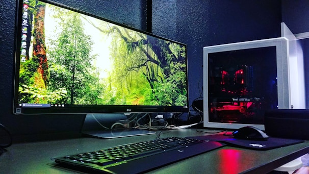 A sleek computer setup featuring a large monitor displaying a forest scene with vibrant greenery. Next to the monitor is a white tower with visible red lighting from internal components. A keyboard and mouse are placed in front, on a desk with a dark surface.