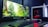 A sleek computer setup featuring a large monitor displaying a forest scene with vibrant greenery. Next to the monitor is a white tower with visible red lighting from internal components. A keyboard and mouse are placed in front, on a desk with a dark surface.