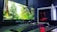 A sleek computer setup featuring a large monitor displaying a forest scene with vibrant greenery. Next to the monitor is a white tower with visible red lighting from internal components. A keyboard and mouse are placed in front, on a desk with a dark surface.