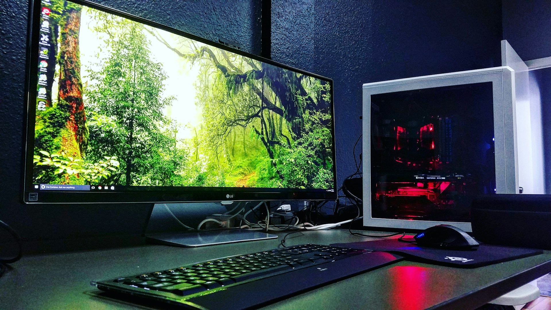 A sleek computer setup featuring a large monitor displaying a forest scene with vibrant greenery. Next to the monitor is a white tower with visible red lighting from internal components. A keyboard and mouse are placed in front, on a desk with a dark surface.