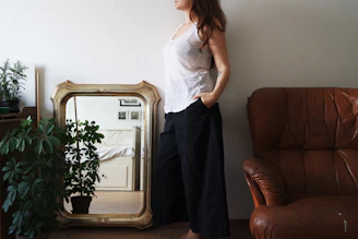 A person stands next to a large, ornate mirror leaning against the wall. The mirror reflects parts of a room featuring a bed and wall decorations. Potted plants are placed on the left, and a brown leather chair is on the right. The person is wearing a white sleeveless top and dark pants.