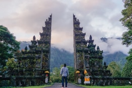 A person stands in front of a traditional Balinese split gate surrounded by lush greenery. The intricate stone architecture is adorned with moss and vegetation, and the background features mist-covered mountains and clouds.