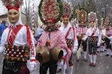 A group of people wearing ornate traditional costumes featuring masks, colorful embroidered shirts, and belts adorned with bells. They are participating in what appears to be a festive parade, with intricate headdresses decorated with wreaths and ornaments. Snow falls gently, adding a wintry atmosphere to the scene.