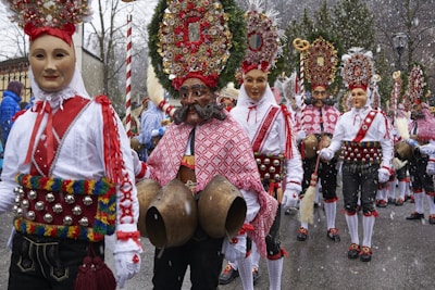 A group of people wearing ornate traditional costumes featuring masks, colorful embroidered shirts, and belts adorned with bells. They are participating in what appears to be a festive parade, with intricate headdresses decorated with wreaths and ornaments. Snow falls gently, adding a wintry atmosphere to the scene.