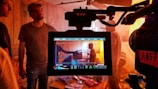 A film set is captured with a focus on a camera monitor displaying a scene being filmed. Multiple people are present, some in the background and one prominently in the center of the monitor, possibly holding a clapperboard. The lighting is warm with a slightly cluttered environment including chairs and various items.