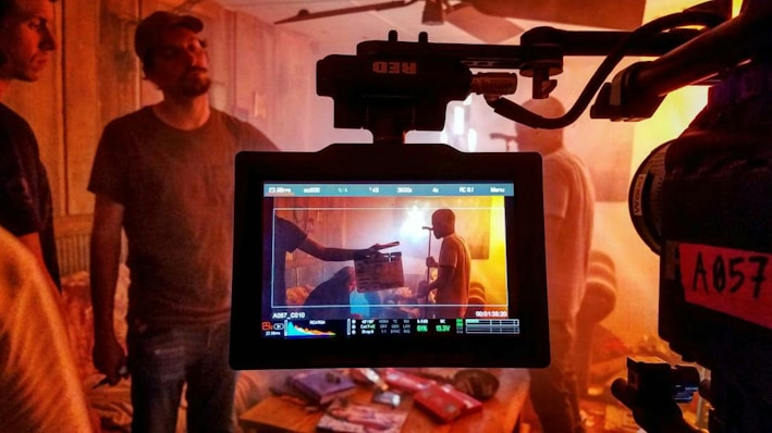 A film set is captured with a focus on a camera monitor displaying a scene being filmed. Multiple people are present, some in the background and one prominently in the center of the monitor, possibly holding a clapperboard. The lighting is warm with a slightly cluttered environment including chairs and various items.