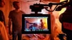A film set is captured with a focus on a camera monitor displaying a scene being filmed. Multiple people are present, some in the background and one prominently in the center of the monitor, possibly holding a clapperboard. The lighting is warm with a slightly cluttered environment including chairs and various items.
