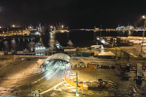 A nighttime view of a bustling port area, illuminated by streetlights and building lights. The scene includes a harbor filled with various docked boats and ships, an array of shipping containers, and crisscrossing railway tracks. The waterfront is flanked by industrial buildings and cranes, contributing to a busy maritime atmosphere.