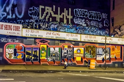 A vibrant mural on a city wall featuring large, colorful letters spelling out 'CHINATOWN'. Each letter contains elements and symbols representative of Chinese culture and urban life. The background includes various graffiti and tags, adding to the urban aesthetic. Above the mural, there are signs in Chinese characters promoting different foods like snow pea, broccoli, and napa. Traffic cones and a construction barrier can be seen in front of the mural.